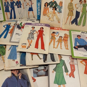 May include: A collection of vintage Simplicity sewing pattern envelopes featuring various clothing designs including dresses, trouser suits, blouses, and swimwear. The paper envelopes display illustrations of people wearing the garments with printed text and numbers.
