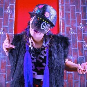 May include: A person wearing a black baseball cap with patches and the word "MAGE" on a face covering. They are wearing a black fur vest, a black and white graphic print shirt, and purple braids. The background is a brick wall.