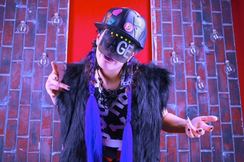 May include: A person wearing a black baseball cap with patches and the word "MAGE" on a face covering. They are wearing a black fur vest, a black and white graphic print shirt, and purple braids. The background is a brick wall.