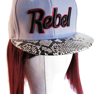 Op de afbeelding: Lichtblauwe baseballcap met het woord "Rebel" in zwart geborduurd met rode rand. De klep heeft een zwart-wit slangenpatroon. De pet wordt getoond op een witte mannequinhoofd met lang, donkerrood haar.