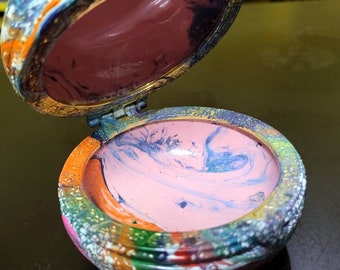 Può includere: Una scatola rotonda e colorata per piccoli oggetti con un design astratto a spirale. La scatola è aperta, rivelando un interno rosa con un motivo a spirale blu e bianco.