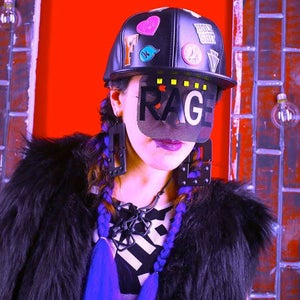 Pode incluir: Boné de beisebol preto com patches e a palavra "RAGE" em uma viseira de malha. A pessoa está usando brincos pretos grandes, um casaco de pele preto e tem tranças roxas. O fundo é uma parede de tijolos com lâmpadas.