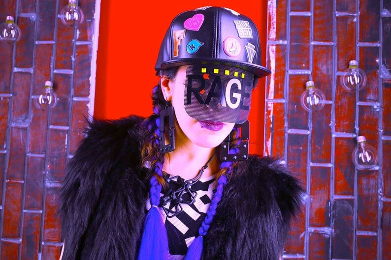 May include: A black baseball cap with patches and the word "RAGE" on a mesh visor. The person is wearing large black earrings, a black fur coat, and has purple braids. The background is a brick wall with light bulbs.