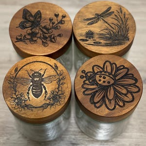 May include: Four glass jars with wooden lids. Each lid features a different nature-themed design: a butterfly with flowers, a dragonfly over water, a bee surrounded by a floral wreath, and a ladybug on a daisy.