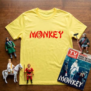 May include: A yellow t-shirt with the word "MONKEY" in red, along with several figurines and a TV Guide magazine. The magazine cover features the word "MONKEY" and images of characters.