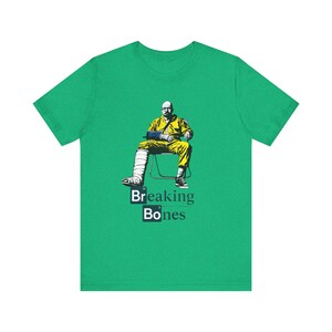 Puede incluir: Camiseta verde con un gráfico de un hombre con un traje amarillo, con un brazo y una pierna rotos. El texto "Breaking Bones" está debajo de la imagen, con "Breaking" encima de "Bones".