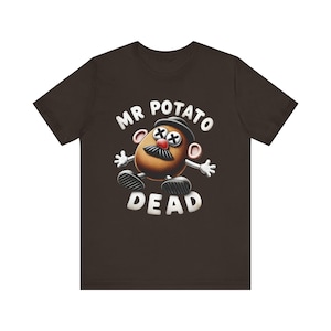 May include: Dark brown t-shirt with a cartoon Mr. Potato Head design. The character has crossed-out eyes, a mustache, and the word "DEAD" below. The text "MR POTATO" is arched above the character in white.