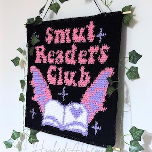 Może przedstawiać: Czarny szydełkowy obraz na ścianę z różowym napisem "Smut Readers Club". Poniżej tekstu znajduje się nietoperz z różowymi i fioletowymi skrzydłami nad otwartą książką. Ozdobiony sztucznym bluszczem.