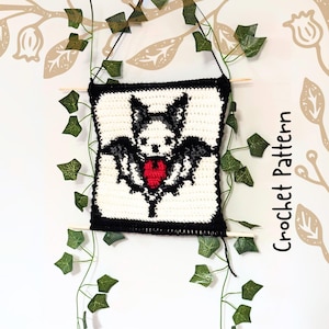 May include: A square, crocheted wall hanging with a bat design and a red heart. The bat has black wings and a white face, set against a white background with a black border. The text "Crochet Pattern" is visible. It is suspended by a wooden dowel.