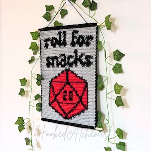 Könnte beinhalten: Gehäkeltes Wandbehang mit dem Text "roll for snacks" über einem roten und schwarzen D20-Würfel mit der Zahl 20. Das Banner ist grau mit schwarzem Rand und hängt an einer Wand mit grünem Efeu.