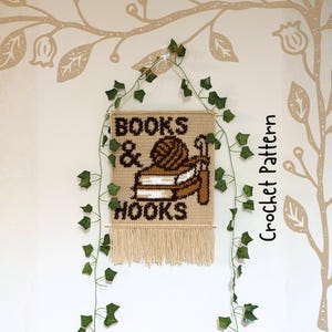 May include: A beige crochet wall hanging with the words "BOOKS & HOOKS" in brown. The design includes a yarn ball, books, and a crochet hook. The bottom has a fringe. Green vines and a tree design surround the sign.