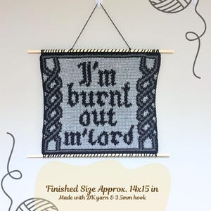 Op de afbeelding: Een handgemaakte, grijs en zwart gehaakte wandhanger met de tekst "I'm burnt out m'lord". De afmeting is ongeveer 35,5 x 38 cm. Opgeschort aan een houten stok met zwart touw.