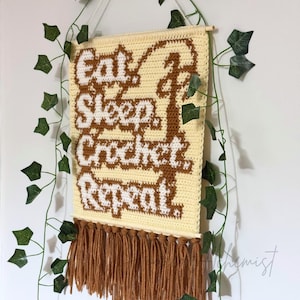 May include: A cream-colored, crocheted wall hanging with the words "Eat Sleep Crochet Repeat" in brown and white. The piece has a fringe of brown yarn at the bottom and is suspended by a wooden dowel and white string. Green ivy vines frame the piece.