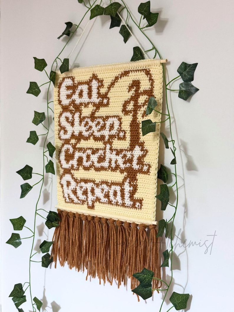 May include: A cream-colored, crocheted wall hanging with the words "Eat Sleep Crochet Repeat" in brown and white. The piece has a fringe of brown yarn at the bottom and is suspended by a wooden dowel and white string. Green ivy vines frame the piece.