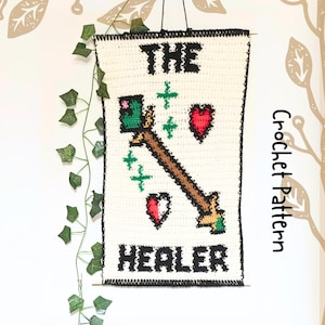 May include: A handmade crochet wall hanging with the words "THE HEALER" in black block letters. The design includes a brown staff with green and gold accents, red hearts, and green crosses on a white background. The words "Crochet Pattern" are written vertically on the right side.