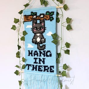 Puede incluir: Un tapiz de pared de ganchillo azul claro con un gato gris colgando de una rama marrón con hojas verdes. Las palabras "HANG IN THERE" están cosidas en negro. La parte inferior tiene flecos. Una clavija de madera y hiedra artificial completan el diseño.