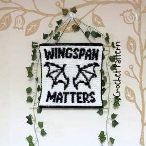 K&ouml;nnte beinhalten: Ein wei&szlig;-schwarzer Wandbehang aus H&auml;kelarbeit mit den Worten "WINGSPAN MATTERS" und einem Fledermausfl&uuml;gel-Design. Das Schild ist mit schwarzem Garn umrandet und mit einer schwarzen Schnur und Holzst&auml;ben aufgeh&auml;ngt. Gr&uuml;ner Efeu schm&uuml;ckt die Wand.