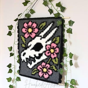 Peut inclure: Suspension murale crochetée avec un motif de crâne blanc, des fleurs roses et des feuilles vertes sur fond gris. Bordure noire festonnée et suspendue à une tringle en bois. Le texte "Hooked Alchemy" est visible.