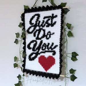 May include: A handmade crochet wall hanging with the words "Just Do You" in black script on a white background. A red heart is below the text. The piece is framed with a black scalloped border and hung with a wooden dowel.