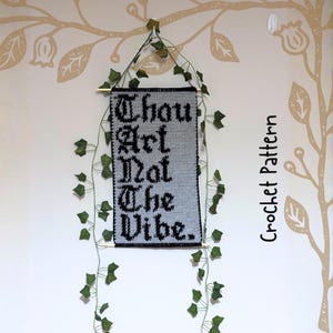Op de afbeelding: Een gehaakte wandhanger met de tekst "Thou Art Not The Vibe" in zwart op een grijze achtergrond. De banner is omlijst met een zwarte rand en versierd met kunst-klimop. De woorden "Crochet Pattern" staan verticaal rechts.