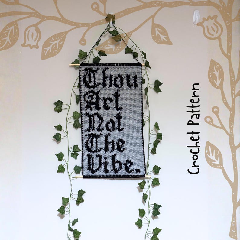 Op de afbeelding: Een gehaakte wandhanger met de tekst "Thou Art Not The Vibe" in zwart op een grijze achtergrond. De banner is omlijst met een zwarte rand en versierd met kunst-klimop. De woorden "Crochet Pattern" staan verticaal rechts.