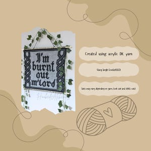 Op de afbeelding: Een zwart en grijs gehaakte wandhanger met de tekst "I'm burnt out m'lord". Het stuk is omlijst met een donkergrijze rand en opgehangen met een houten deuvel. Groene klimopachtige ranken zijn rond het stuk gedrapeerd. De tekst "Hooked Alchemy" staat onderaan.
