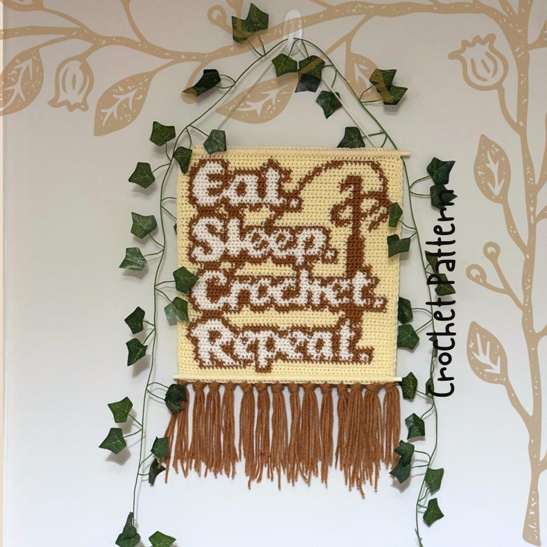 May include: A crocheted wall hanging with the words "Eat Sleep Crochet Repeat" in brown and white. The banner is cream-colored with brown fringe at the bottom. Green ivy and a decorative wall pattern surround the banner.