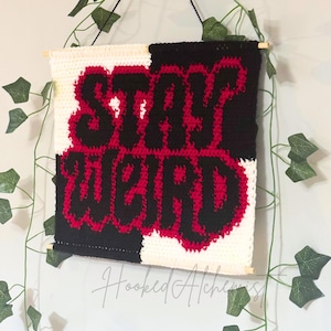 Op de afbeelding: Een handgemaakte gehaakte wandhanger met de woorden "STAY WEIRD" in rood en zwart. De achtergrond is verdeeld in zwarte en witte secties. Het stuk is opgehangen met een houten deuvel en zwart touw.