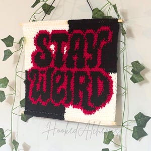 May include: A handmade crochet wall hanging with the words "STAY WEIRD" in red and black. The background is split into black and white sections. The piece is hung with a wooden dowel and black string.