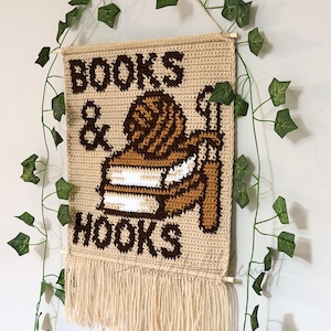 May include: A beige crochet wall hanging with the words "BOOKS & HOOKS" in brown. The design includes a yarn ball, books, and crochet hooks. The bottom edge has a fringe, and it hangs from a wooden dowel.