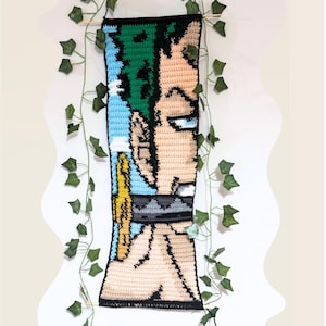 May include: Crocheted wall hanging featuring a character with green hair and a blue eye. The artwork is in shades of green, blue, tan, and black. The piece is rectangular and hangs from a wooden dowel, with faux ivy vines around it.