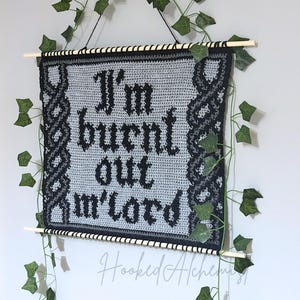 以下が含まれることがあります： 黒いゴシック体で「I'm burnt out m'lord」と書かれた灰色の手編み壁掛け。黒とグレーのケルトノットのデザインで縁取られています。木製の棒と人工のツタが装飾を完成させています。