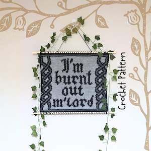 Op de afbeelding: Een gehaakte wandhanger met de tekst "I'm burnt out m'lord" in zwarte gotische letters. De grijze banner heeft een zwarte rand en decoratieve accenten. Bovenop een houten deuvel en een ophangtouw. Groene wijnranken en bladeren zijn gedrapeerd.