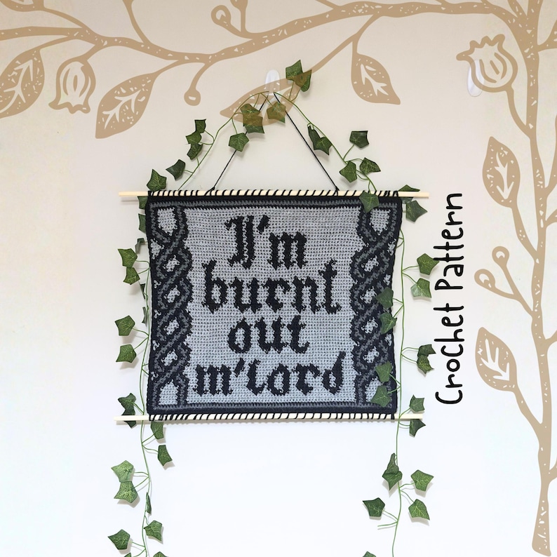 Op de afbeelding: Een gehaakte wandhanger met de tekst "I'm burnt out m'lord" in zwarte gotische letters. De grijze banner heeft een zwarte rand en decoratieve accenten. Bovenop een houten deuvel en een ophangtouw. Groene wijnranken en bladeren zijn gedrapeerd.