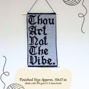 Op de afbeelding: Een handgemaakte, lichtgrijze gehaakte wandhanger met de tekst "Thou Art Not The Vibe" in zwarte gotische letters. De afmeting is ongeveer 25 x 43 cm. Het is opgehangen aan een houten stok met zwart touw.