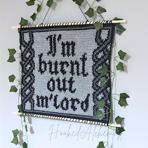 Puede incluir: Un tapiz de pared de ganchillo gris con la frase "I'm burnt out m'lord" en letras góticas negras. El fondo está enmarcado con un diseño de nudos celtas negros y grises. Varillas de madera y hiedra artificial completan la decoración.