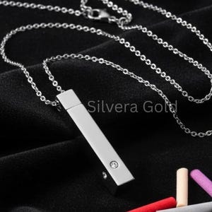 May include: A silver-colored necklace with a rectangular pendant and a small, clear gemstone. The necklace is displayed on a black surface, with a collection of colorful, cylindrical objects nearby. The text "Silvera Gold" is visible.