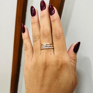 May include: A stack of three rose gold rings worn on a finger. The top ring features a textured honeycomb pattern, the middle ring has a smooth band, and the bottom ring is set with a row of small, round, clear stones along the entire circumference.