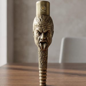 May include: A bronze-colored bottle stopper with a detailed, carved face design. The face has an open mouth with a protruding tongue and is topped with a cylindrical cap. The text "SilverGold" is visible on the cap. The stopper is likely for wine or spirits.