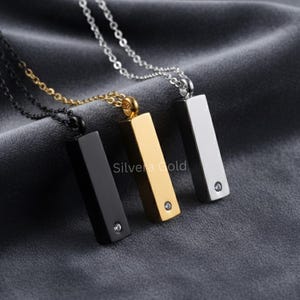 Op de afbeelding: Drie rechthoekige hangerkettingen in zwart, goud en zilver. Elke hanger heeft een kleine, heldere edelsteen en hangt aan een delicate ketting. De tekst "Silvera Gold" is zichtbaar op de gouden hanger.