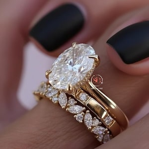 Puede incluir: Un juego de anillos de oro de tres piezas. El anillo de compromiso presenta un gran diamante de talla ovalada con un engaste de halo. La alianza tiene diamantes talla marquesa. La tercera banda es una banda de oro simple.