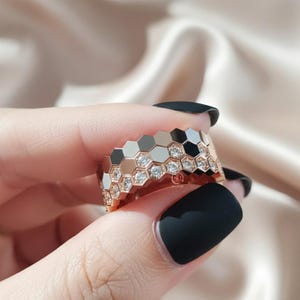 Può includere: Anello in oro rosa con design a nido d'ape. L'anello presenta forme esagonali nere, bianche e oro rosa alternate, con piccole pietre preziose trasparenti incastonate al centro di alcuni esagoni.