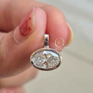 May include: A silver-toned pendant featuring a clear, oval-cut gemstone set in a smooth bezel setting. The pendant includes a small vertical bail at the top for attachment. A circular logo with the text Silvera Gold is visible on the right side.
