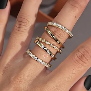Könnte beinhalten: Eine Auswahl an goldenen Ringen, jeweils mit einem einzigartigen Design. Ein Ring hat ein Band aus kleinen, funkelnden Steinen. Andere Ringe haben eine glatte, polierte Oberfläche oder sind mit zarten, edelsteinartigen Akzenten verziert. Die Ringe werden an einem Finger mit schwarzem Nagellack präsentiert.
