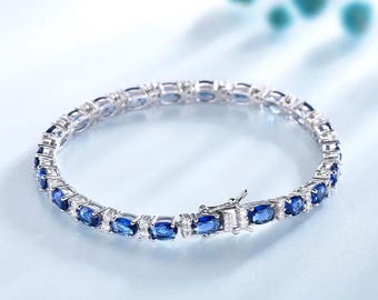 May include: A silver bracelet featuring oval-cut blue gemstones and small white stones. The bracelet has a secure clasp and is presented on a light blue surface. The jewellery is elegant and suitable for special occasions.