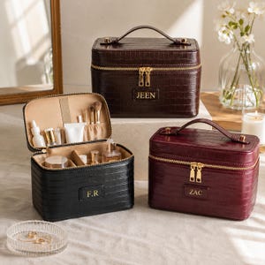 May include: Three rectangular cosmetic cases with a crocodile-embossed texture, gold-tone zippers, and top handles. One case is open, revealing beige interior compartments with various beauty products. Each case features a personalized name tag.