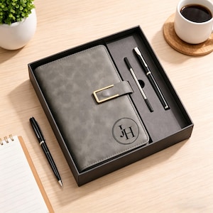 May include: A gray journal with a gold clasp and the initials "JH" in a circle, presented in a black box with two black pens. The journal is closed and has a textured surface. A cup of coffee and a small plant are also visible.