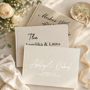 Könnte beinhalten: Drei cremefarbene Hochzeitsgästebücher mit individuellem Text. Das obere Buch trägt die Aufschrift "Mesher" mit floralen Akzenten. Das mittlere Buch lautet "The Angelika & Laura". Das untere Buch lautet "Ashley & Cabral" mit Datum und Ort.