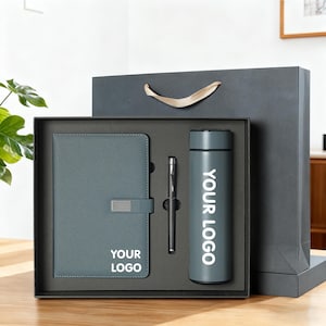 May include: A corporate gift set in a black box, including a grey notebook with a silver clasp, a black pen, and a grey insulated flask. The flask and notebook have the text "YOUR LOGO" printed in white. A grey gift bag is behind the box.