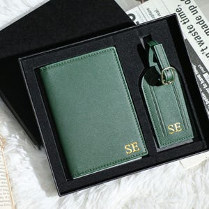 以下が含まれることがあります： 金色のイニシャル「SE」が入った緑色のパスポートホルダーとラゲッジタグのセット。黒いギフトボックスに入っています。セットはテクスチャード加工された素材で作られています。ラゲッジタグにはバックルとストラップが付いています。